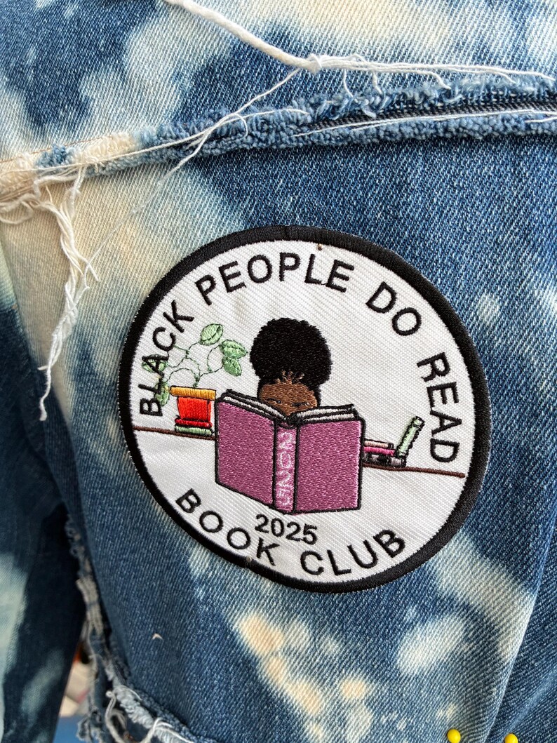May include: A denim jacket with a circular patch showing the text "BLACK PEOPLE DO READ BOOK CLUB 2025" around an illustration of a person reading. The jacket has a bleached, distressed design, with visible frayed edges.