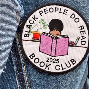 May include: A circular patch featuring the text "BLACK PEOPLE DO READ BOOK CLUB 2025" around an illustration of a person reading a pink book. A small potted plant and a pen are also shown. The patch is set against a denim backdrop.