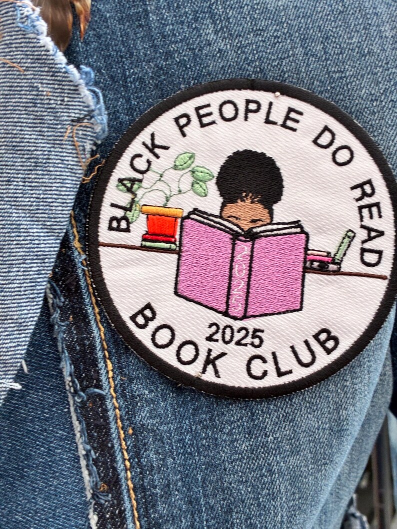 May include: A circular patch featuring the text "BLACK PEOPLE DO READ BOOK CLUB 2025" around an illustration of a person reading a pink book. A small potted plant and a pen are also shown. The patch is set against a denim backdrop.