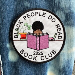 May include: Circular embroidered patch with the text "BLACK PEOPLE DO READ BOOK CLUB 2025". The design includes a black person reading a pink book, a potted plant, and a pencil, all within a black border.