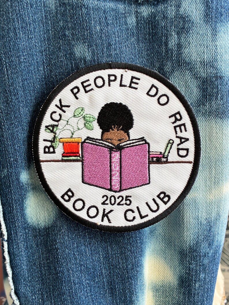 May include: Circular embroidered patch with the text "BLACK PEOPLE DO READ BOOK CLUB 2025". The design includes a black person reading a pink book, a potted plant, and a pencil, all within a black border.