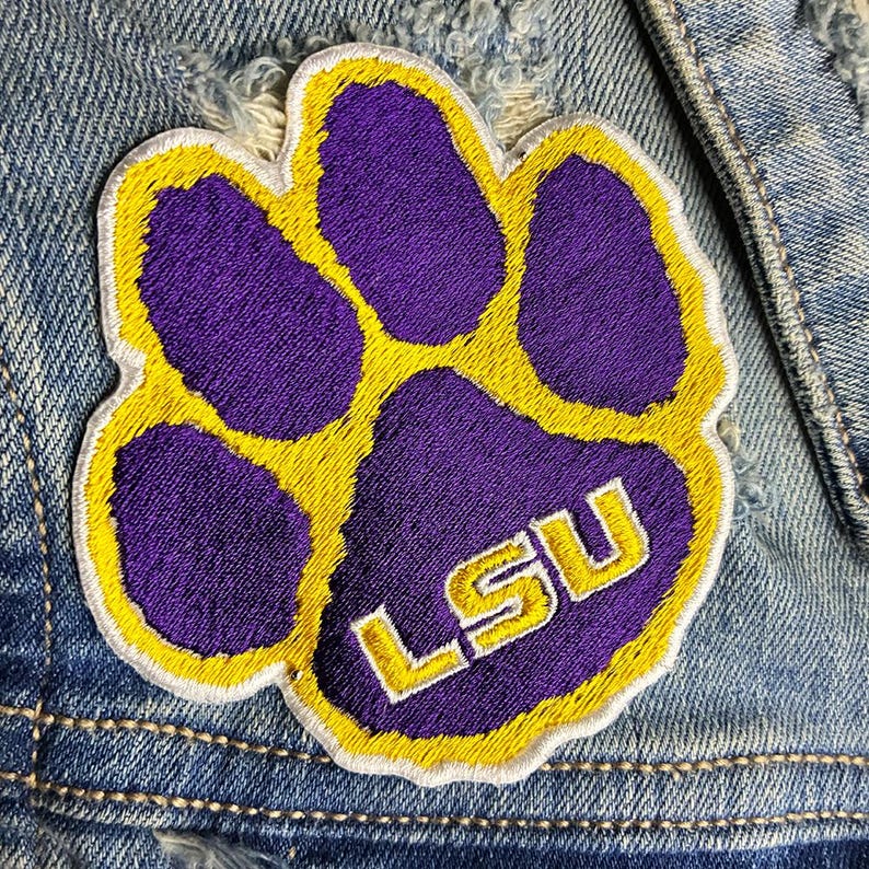 Peut inclure: &Eacute;cusson brod&eacute; en forme de patte de couleur violette et dor&eacute;e avec les lettres "LSU" en or. L'&eacute;cusson est cousu sur une veste en jean d&eacute;lav&eacute;.