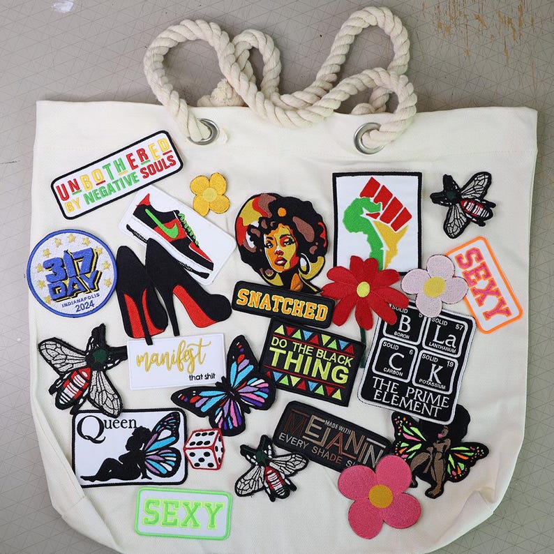 Peut inclure: Un sac fourre-tout cr&egrave;me avec une poign&eacute;e en corde &eacute;paisse, orn&eacute; de divers &eacute;cussons brod&eacute;s. Les &eacute;cussons comprennent des papillons, des fleurs, des talons hauts, des baskets et des textes comme "Unbothered" et "Snatched."