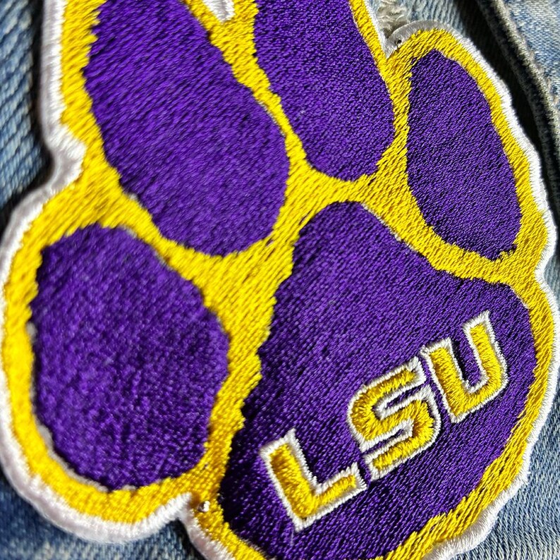 Peut inclure: &Eacute;cusson brod&eacute; repr&eacute;sentant une empreinte de patte violette avec des accents jaunes et blancs. Les lettres "LSU" sont brod&eacute;es en jaune avec des contours blancs. L'&eacute;cusson est probablement destin&eacute; aux v&ecirc;tements ou aux accessoires.