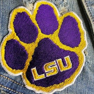 Peut inclure: &Eacute;cusson brod&eacute; en forme d'empreinte de patte violette avec une bordure jaune et les lettres "LSU" en blanc. L'&eacute;cusson est cousu sur une veste en jean.