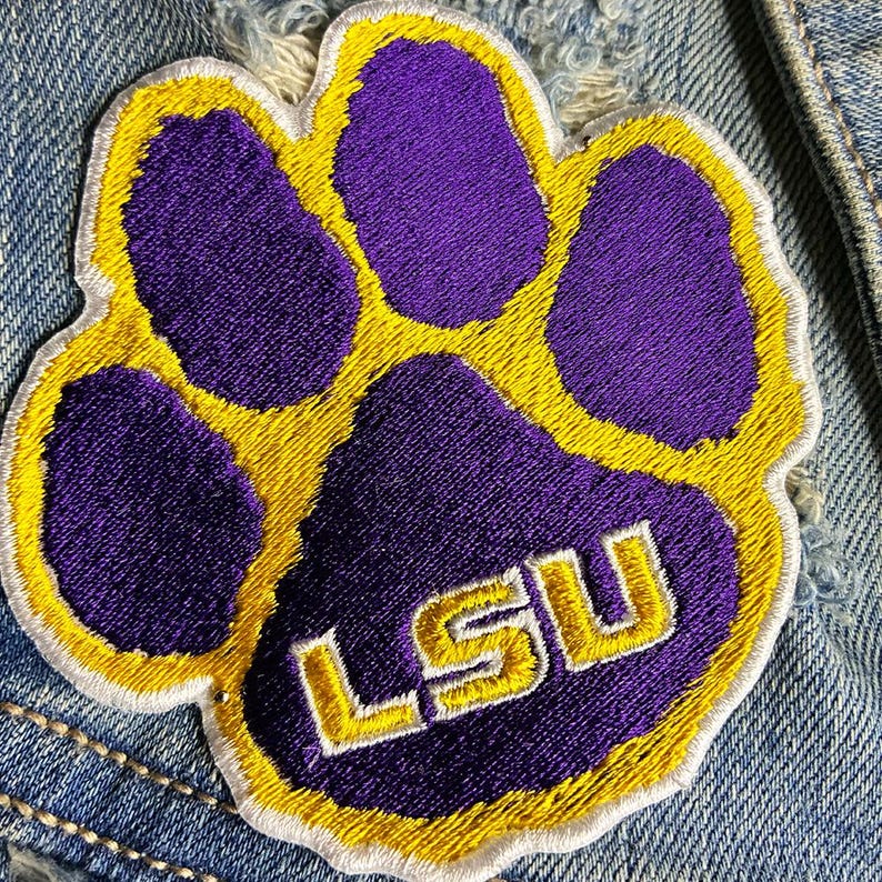Peut inclure: &Eacute;cusson brod&eacute; en forme d'empreinte de patte violette avec une bordure jaune et les lettres "LSU" en blanc. L'&eacute;cusson est cousu sur une veste en jean.