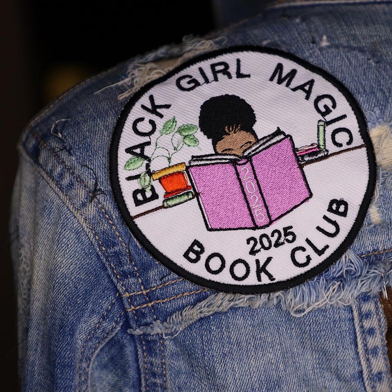 May include: A circular embroidered patch with the text "BLACK GIRL MAGIC BOOK CLUB 2025". The patch features a Black girl reading a pink book, a plant, and makeup items. The patch is attached to a denim jacket.