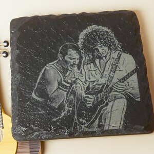May include: Black and white engraved image of two musicians on a black slate coaster. The musician on the left is singing into a microphone and the musician on the right is playing an electric guitar.