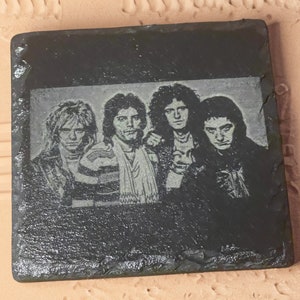 May include: Black and white image of a rock band on a black slate coaster. The band members are all wearing dark clothing and have long hair. The image is a close-up of the band's faces.