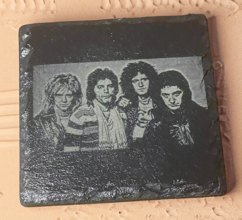May include: Black and white image of a rock band on a black slate coaster. The band members are all wearing dark clothing and have long hair. The image is a close-up of the band's faces.