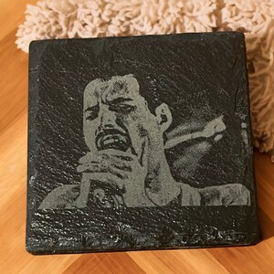 May include: A black slate coaster with a white engraved image of a person singing into a microphone.