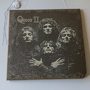 May include: A black and white image of the rock band Queen on a slate plaque. The band members are arranged in a circle with the band's name "Queen II" at the top.