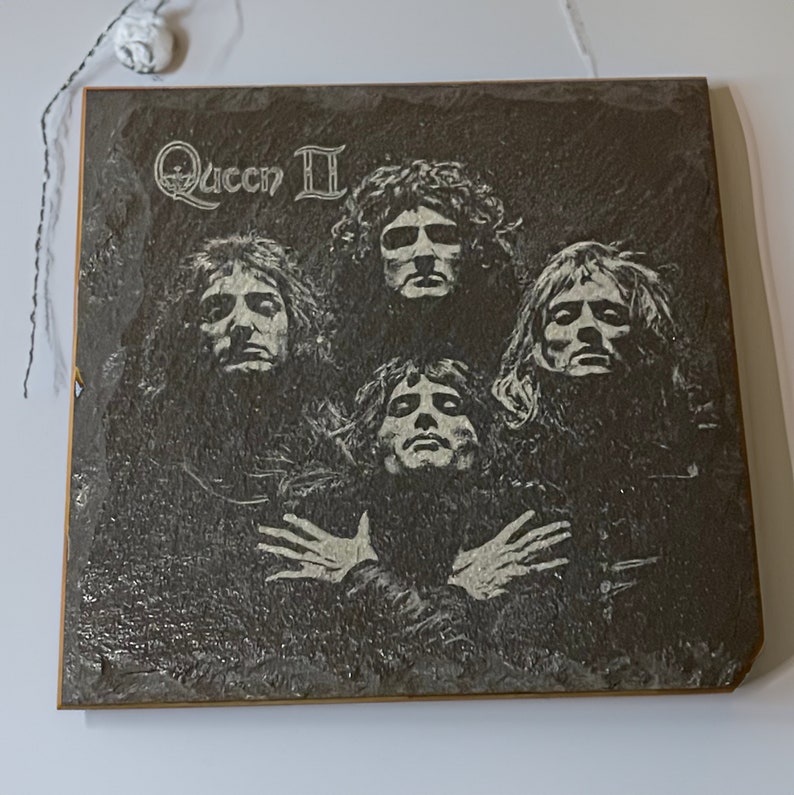 May include: A black and white image of the rock band Queen on a slate plaque. The band members are arranged in a circle with the band's name "Queen II" at the top.