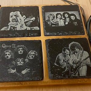 May include: Set of four black slate coasters featuring images of the rock band Queen. The coasters feature images of Freddie Mercury, Brian May, Roger Taylor, and John Deacon. The coasters are arranged in a square shape on a wooden base.