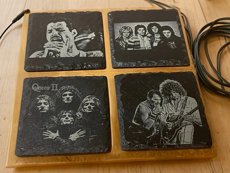 May include: Set of four black slate coasters featuring images of the rock band Queen. The coasters feature images of Freddie Mercury, Brian May, Roger Taylor, and John Deacon. The coasters are arranged in a square shape on a wooden base.