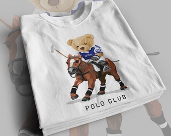 May include: White t-shirt with a graphic of a teddy bear riding a brown horse and holding a polo mallet. The bear is wearing a blue polo shirt with the text "CAPTAIN" and the number "03". The text "POLO CLUB" is printed below the graphic.