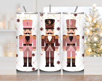 May include: Three stainless steel insulated tumblers featuring a festive nutcracker design. Each tumbler showcases a different nutcracker in shades of pink, brown, and tan, with a black mustache and hat. The background is white with snowflake and heart accents.