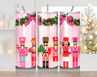 May include: Pink tumbler featuring a festive design with nutcracker soldiers, candy canes, and holly. The tumbler has a pink and white striped background, a pink bow, and a silver straw. A Christmas-themed tumbler.