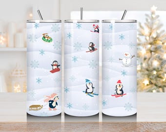 May include: A set of three stainless steel insulated tumblers with a winter theme. Each tumbler features a white background with illustrations of penguins skiing, sledding, and playing in the snow. The tumblers have a silver straw.