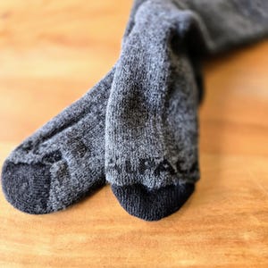 May include: A pair of gray wool socks with black ribbed cuffs. The socks are folded and lying on a wooden surface.