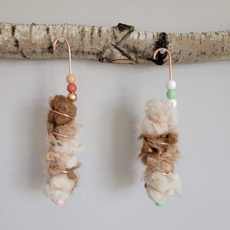 May include: Two brown and white wool pom-poms with copper wire wrapped around them. Each pom-pom is hanging from a copper hook with colorful beads.