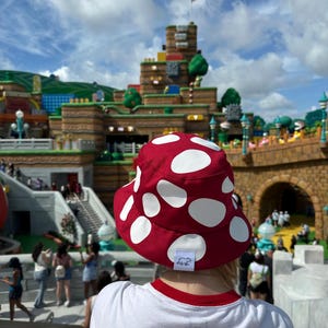 May include: A red bucket hat with large white polka dots. The hat is worn by a person in a white t-shirt with a red trim. The background shows a theme park with a castle-like structure and people.