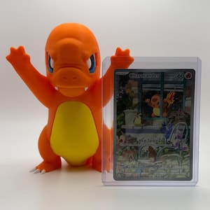 May include: An orange 3D-printed Charmander figure with outstretched arms stands next to a Charmander trading card in a protective sleeve. The card features an illustration of Charmander and the text "Heat Tackle".
