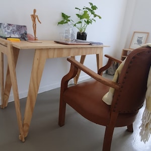 May include: A light-colored wooden desk with angled legs, a brown leather armchair, and a small framed owl print. A plant in a black pot, a wooden artist's mannequin, and a laptop are on the desk. A yellow blanket is draped over the chair.