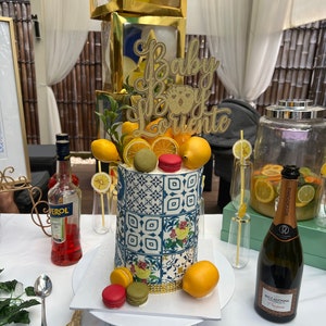 Pu&ograve; includere: Una torta blu e bianca decorata con limoni e macarons. La torta &egrave; sormontata da un cartello dorato con glitter che dice "Baby Boy Lorenzo".