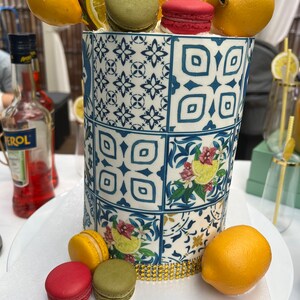 Pu&ograve; includere: Una torta bianca decorata con un motivo a piastrelle blu e bianche, limoni e macarons. La torta &egrave; sormontata da un cartello dorato con glitter che dice "Lemonade".