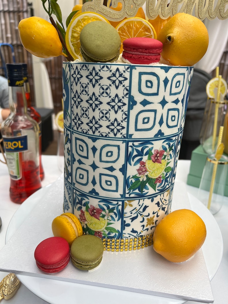 Pu&ograve; includere: Una torta bianca decorata con un motivo a piastrelle blu e bianche, limoni e macarons. La torta &egrave; sormontata da un cartello dorato con glitter che dice "Lemonade".