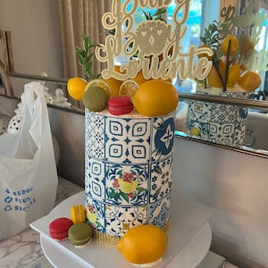 Pu&ograve; includere: Una torta bianca decorata con un motivo a piastrelle blu e bianche, limoni e macarons. La torta &egrave; sormontata da un topper dorato con glitter che dice "Baby Boy Lorenzo".