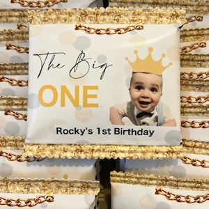 May include: Stacked party favors with a gold chain design. The front favor has the text "The Big ONE" with a photo of a baby wearing a crown. The text "Rocky's 1st Birthday" is also visible.