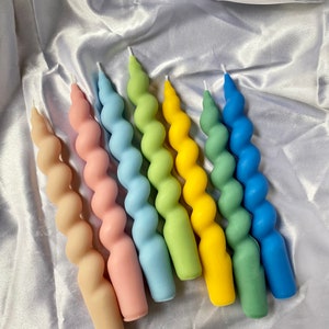 May include: A collection of six spiral-shaped candles in a variety of pastel colors, including beige, pink, light blue, green, yellow, and dark blue. The candles are arranged on a white, satin-like surface.