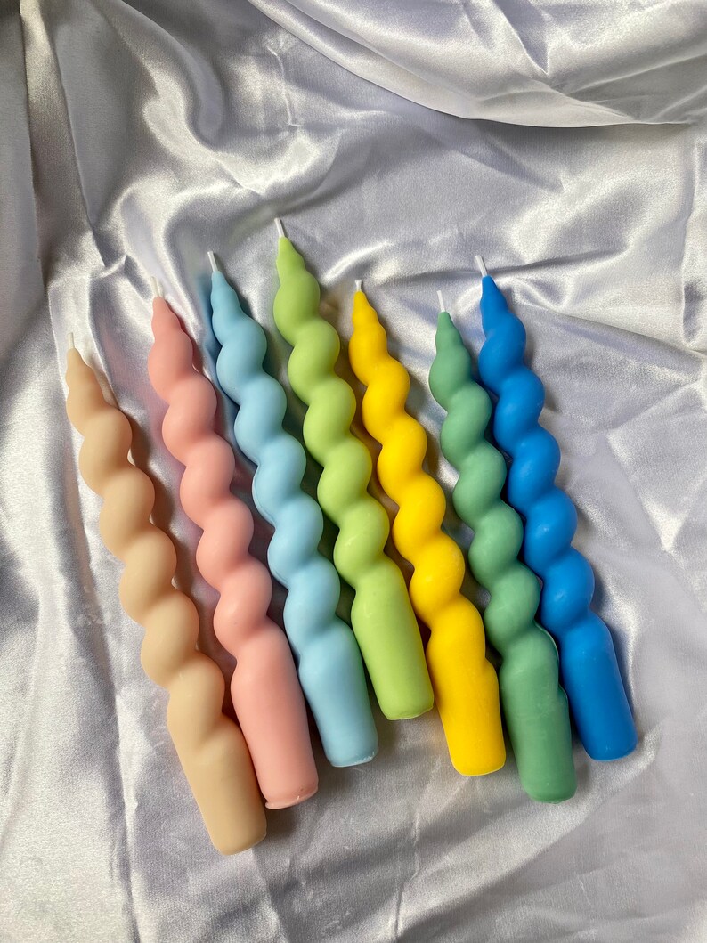 May include: A collection of six spiral-shaped candles in a variety of pastel colors, including beige, pink, light blue, green, yellow, and dark blue. The candles are arranged on a white, satin-like surface.