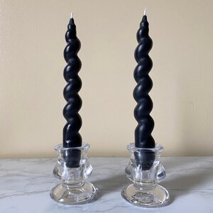 May include: Two black spiral taper candles in clear glass candle holders. The candles have a twisted design and are placed on a white marble surface. The candle holders have a decorative shape.