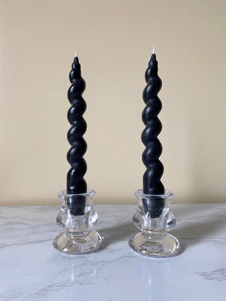May include: Two black spiral taper candles in clear glass candle holders. The candles have a twisted design and are placed on a white marble surface. The candle holders have a decorative shape.