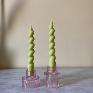 May include: Two green spiral candles sit in pink glass candle holders. The candle holders have a ribbed design and a circular base. The candles are a light green color and have a twisted shape. The background is a neutral color.