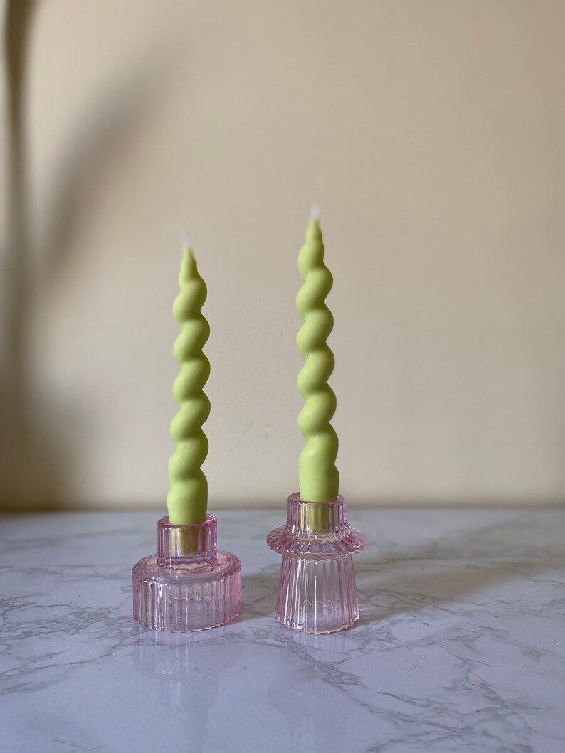 May include: Two green spiral candles sit in pink glass candle holders. The candle holders have a ribbed design and a circular base. The candles are a light green color and have a twisted shape. The background is a neutral color.
