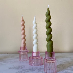 May include: Three spiral candles in pink, white, and green, each set in a pink glass holder. The candles have a twisted design and are of varying heights. The holders have a ribbed texture and a circular base, creating a decorative display.