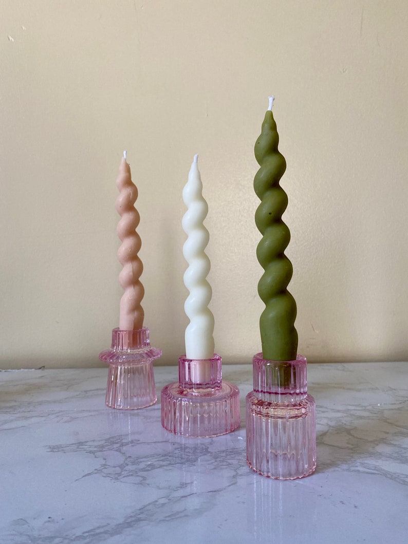 May include: Three spiral candles in pink, white, and green, each set in a pink glass holder. The candles have a twisted design and are of varying heights. The holders have a ribbed texture and a circular base, creating a decorative display.