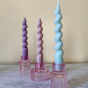 May include: Three spiral candles in shades of purple, pink, and blue, each set in a pink glass holder. The candles have a twisted design and are of varying heights. The holders are a light pink color and have a ribbed texture.