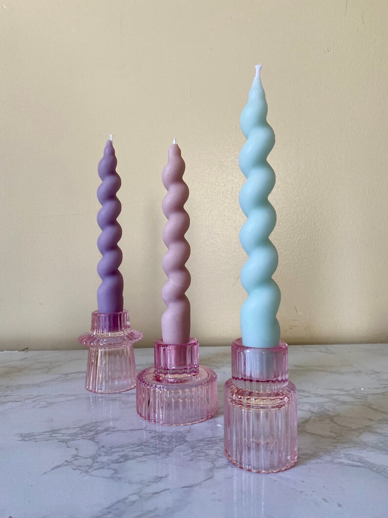 May include: Three spiral candles in shades of purple, pink, and blue, each set in a pink glass holder. The candles have a twisted design and are of varying heights. The holders are a light pink color and have a ribbed texture.