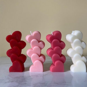May include: Four stacked heart-shaped candles in red, pink, and white. The candles are arranged in a row on a white surface.