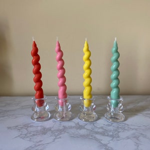 May include: Four colorful spiral candles in red, pink, yellow, and green, each set in a clear glass candle holder. The candles have a twisted design and are arranged in a row on a marble surface.