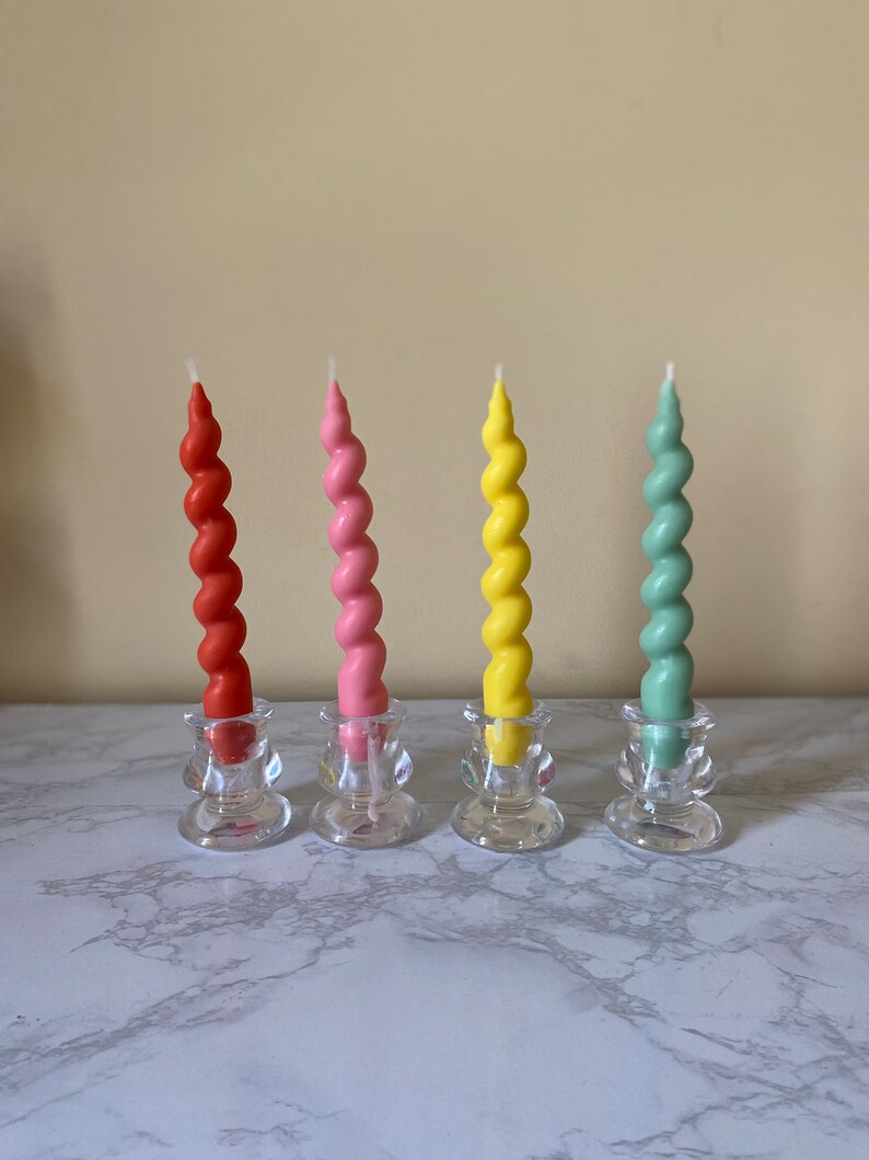 May include: Four colorful spiral candles in red, pink, yellow, and green, each set in a clear glass candle holder. The candles have a twisted design and are arranged in a row on a marble surface.