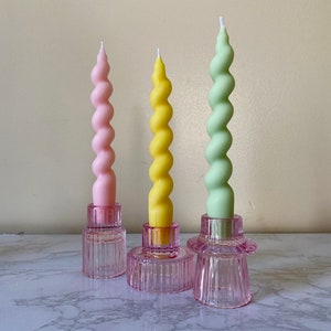 May include: Three spiral candles in pastel colors: pink, yellow, and green. Each candle sits atop a pink glass candle holder. The candles have a twisted design, and the holders have a ribbed texture.