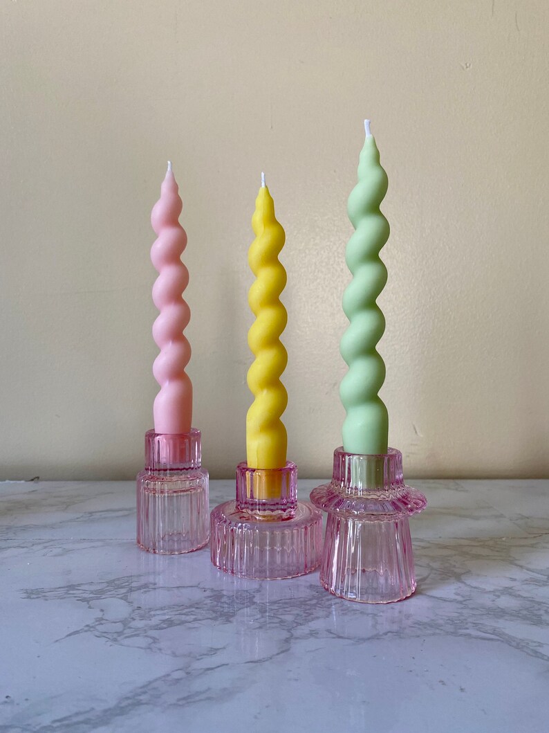 May include: Three spiral candles in pastel colors: pink, yellow, and green. Each candle sits atop a pink glass candle holder. The candles have a twisted design, and the holders have a ribbed texture.