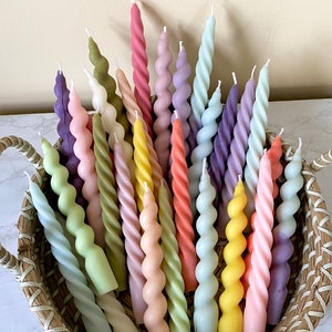 May include: A collection of spiral-shaped candles in a woven basket. The candles are in various pastel colors, including pink, yellow, green, and purple. The candles have a twisted design and are arranged in a decorative display.