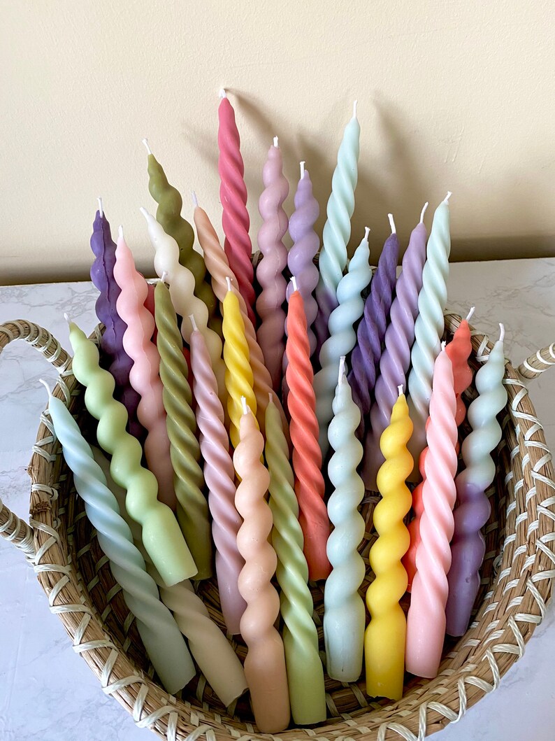 May include: A collection of spiral-shaped candles in a woven basket. The candles are in various pastel colors, including pink, yellow, green, and purple. The candles have a twisted design and are arranged in a decorative display.